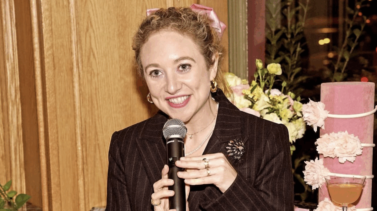 Faran Krentcil in a pinstripe blazer smiling while holding a microphone.